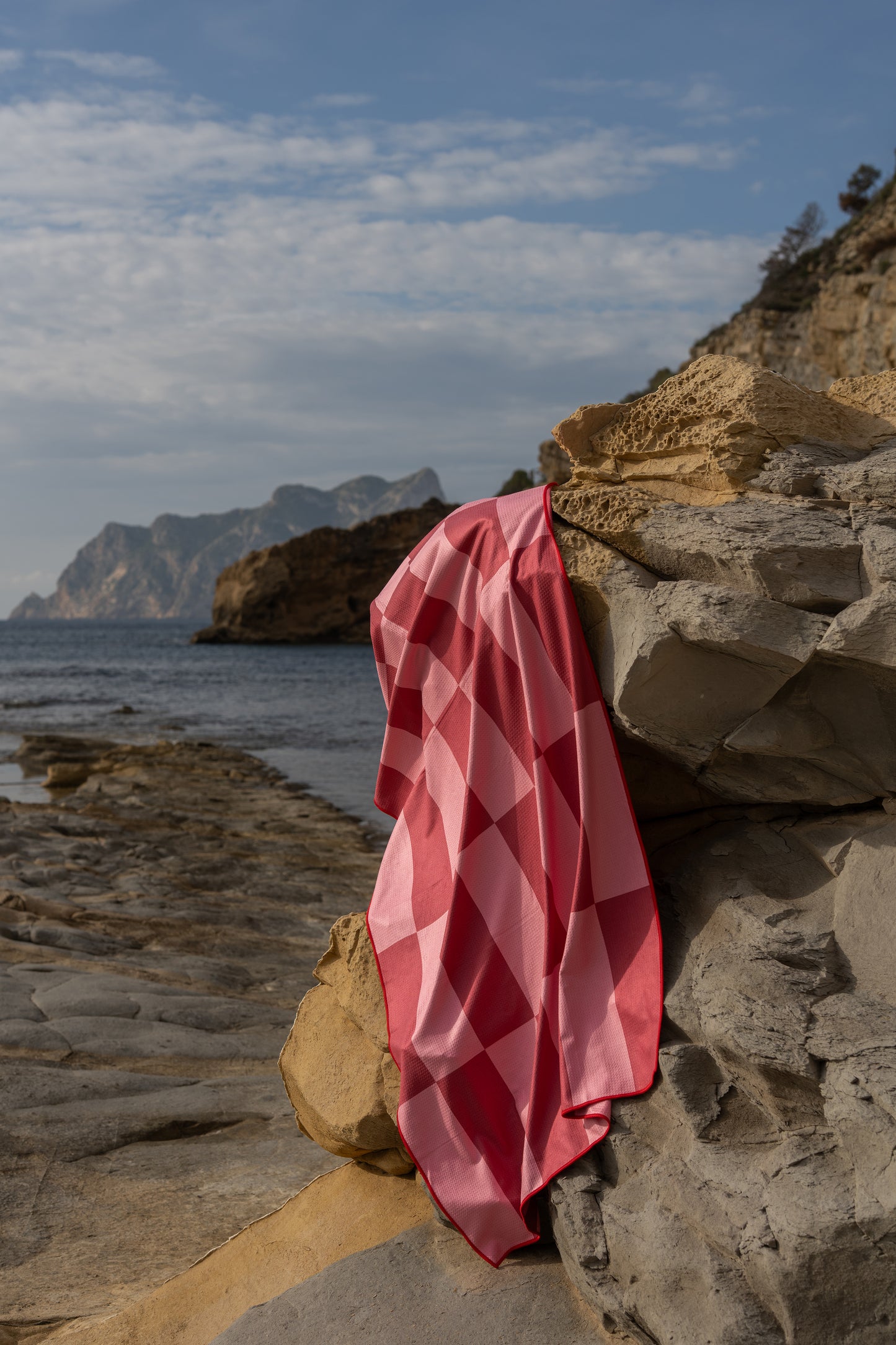 Lichtgewicht beach towel van Piece of Sun in de kleurencombinatie donkerroze met lichtroze, hangend over een rots aan de kustlijn. Gemaakt van microvezel met wafelstructuur en 85% gerecycled materiaal. Perfecte strand metgezel: sneldrogend, compact en stijlvol.