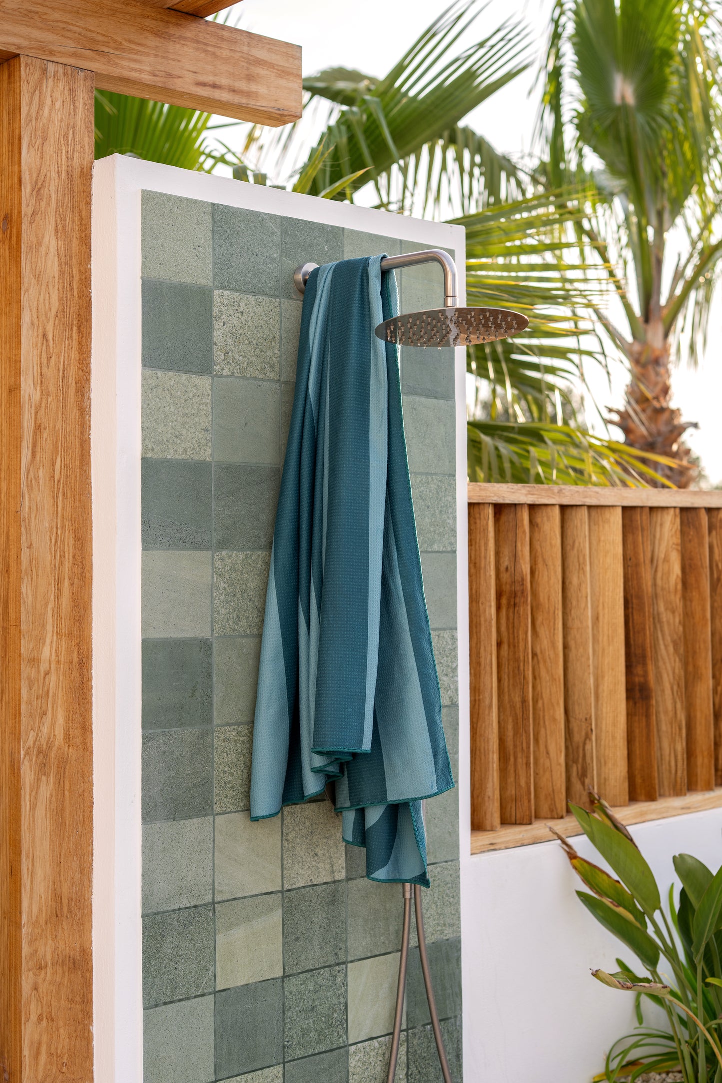 Duurzame beach towel van Piece of Sun in turquoise-petrol kleur hangt aan een buitendouche met groene tegels. Deze strand handdoek is lichtgewicht, zandvrij, sneldrogend en bestaat voor 85% uit gerecycled materiaal – ideaal voor zomerse avonturen.