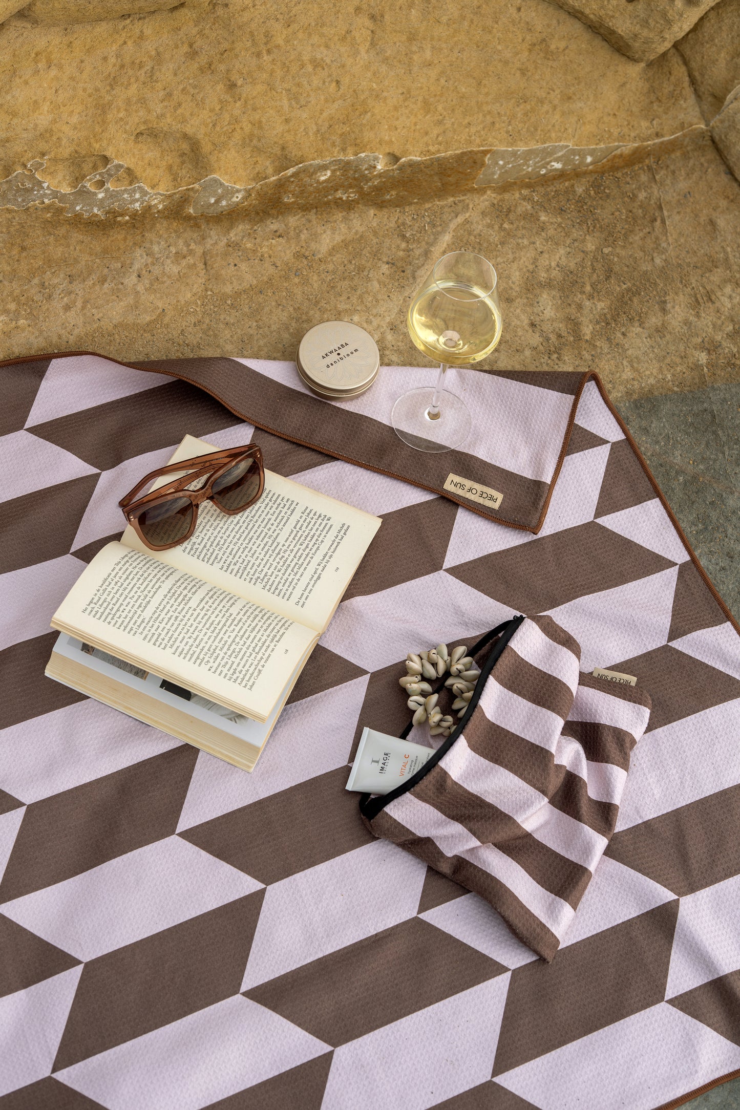 Bruin-beige strandlaken van microvezel met wafelstructuur en geometrisch patroon ligt op een rotsachtige ondergrond. Op het laken liggen een opengeslagen boek met zonnebril, een glas witte wijn, een etui in bijpassend streepjesmotief, schelpenarmband en zonnecrème – klaar voor een ontspannen moment in de zon.