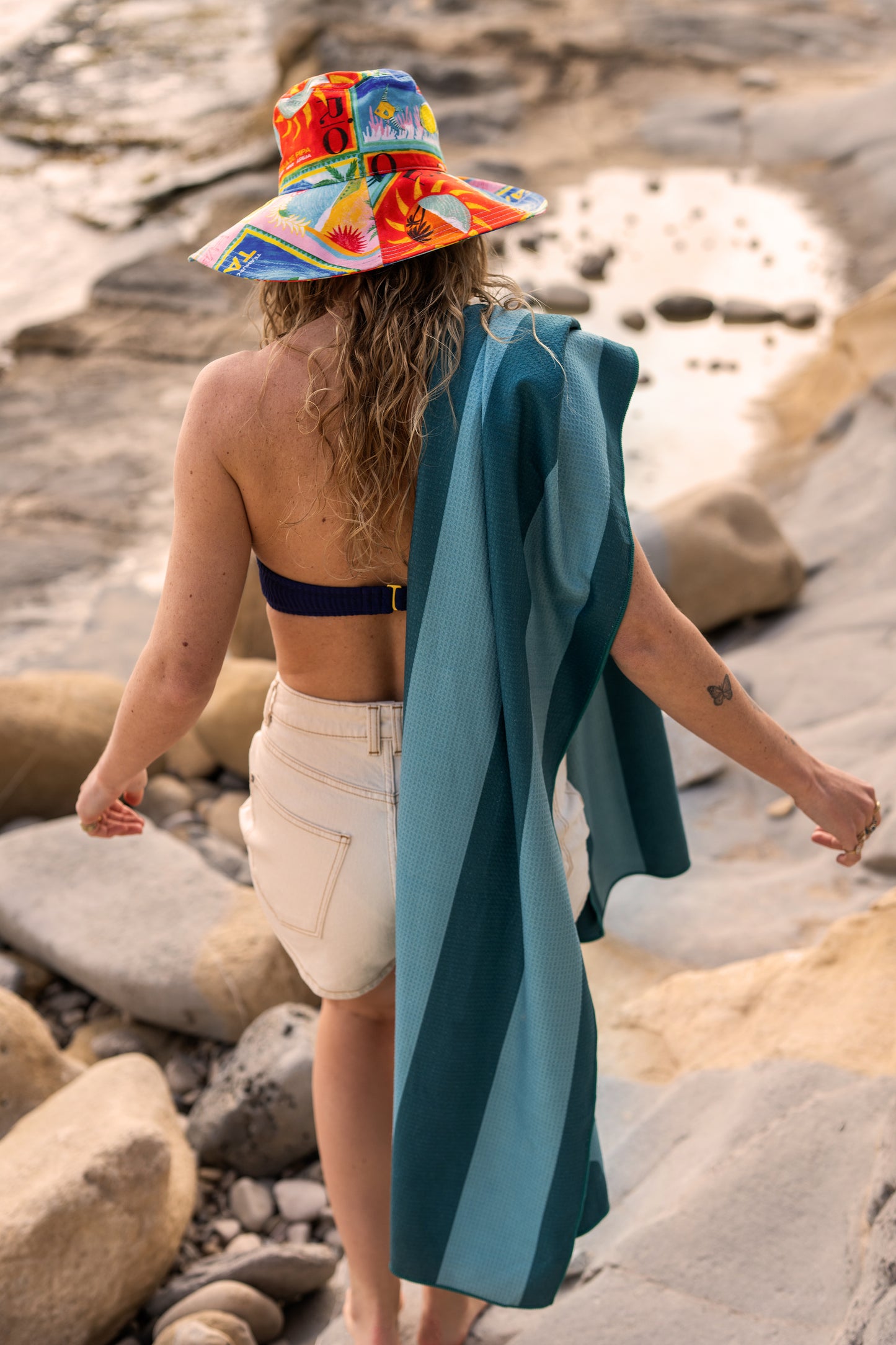 Een vrouw wandelt over rotsen met een turquoise-petrol strandlaken van Piece of Sun losjes over haar schouder. Dit lichtgewicht laken is zandvrij, compact opvouwbaar en gemaakt van 85% gerecycled materiaal.