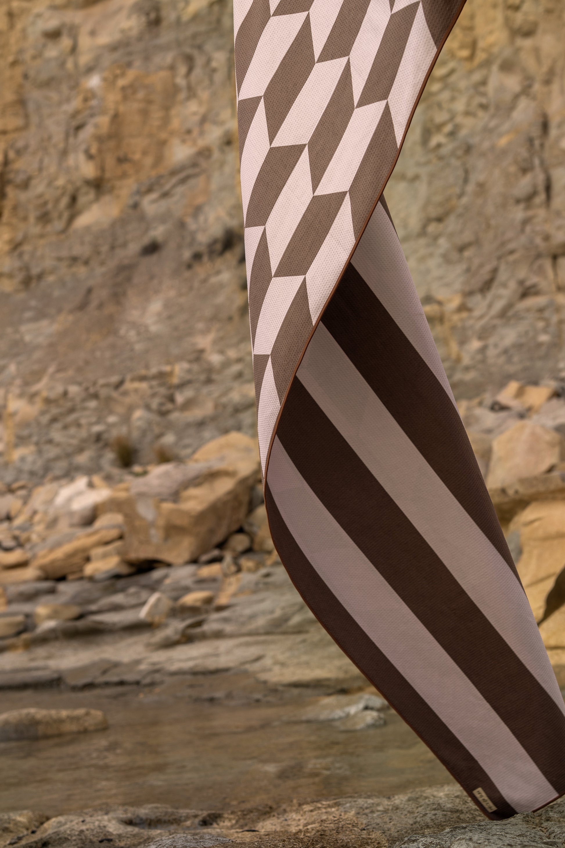 Detailopname van de dubbelzijdige beach towel van Piece of Sun in bruin met beige, met geometrisch patroon aan de ene kant en brede strepen aan de andere. Gemaakt van microvezel met wafelstructuur, zandvrij en sneldrogend. Voor 85% vervaardigd uit gerecycled materiaal.