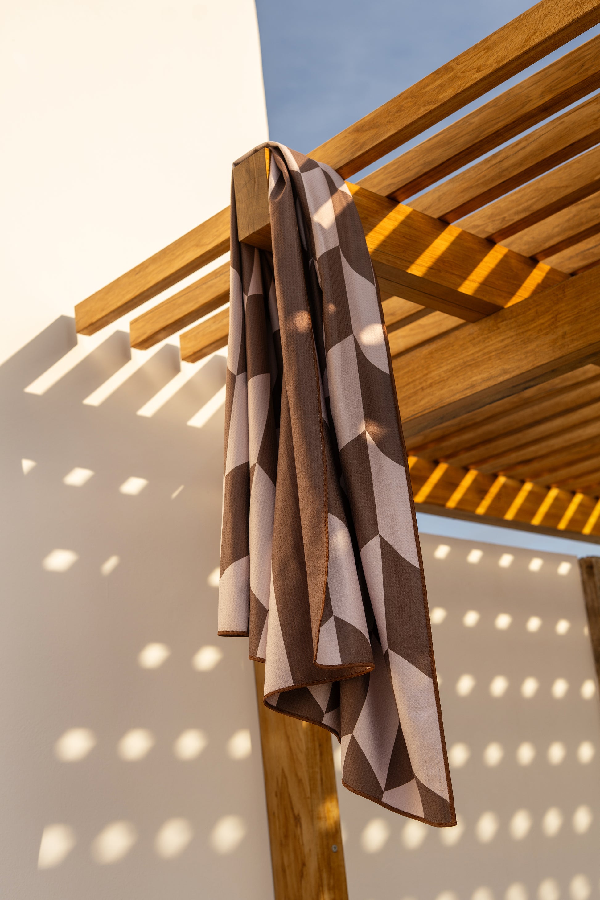 Bruin-beige beach towel van Piece of Sun hangt over een houten pergola in de zon. Het geometrische patroon en de wafelstructuur van de microvezelstof zijn goed zichtbaar. Deze duurzame strand handdoek is lichtgewicht, sneldrogend en voor 85% gemaakt van gerecycled materiaal.