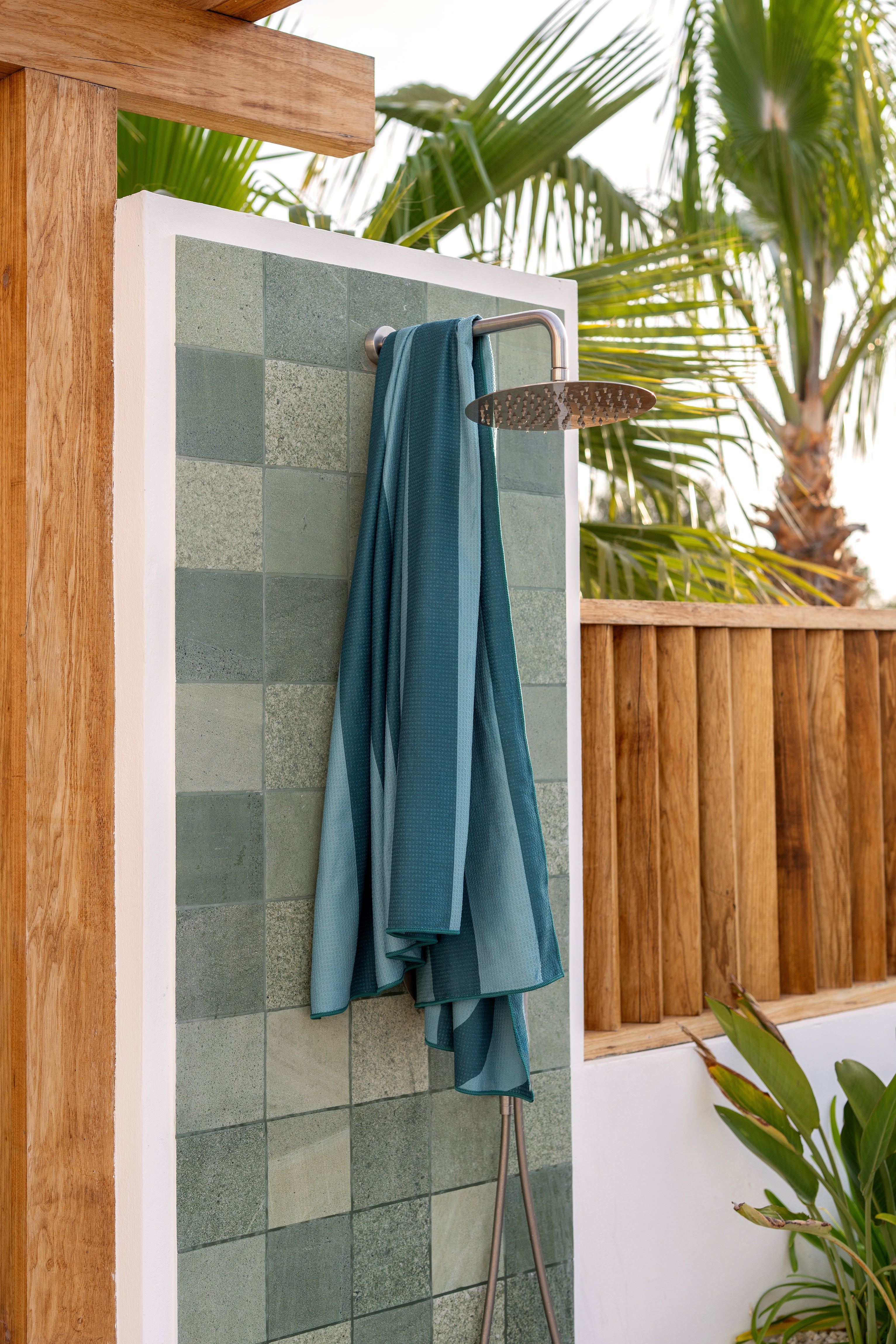 Duurzame beach towel van Piece of Sun in turquoise-petrol kleur hangt aan een buitendouche met groene tegels. Deze strand handdoek is lichtgewicht, zandvrij, sneldrogend en bestaat voor 85% uit gerecycled materiaal – ideaal voor zomerse avonturen.