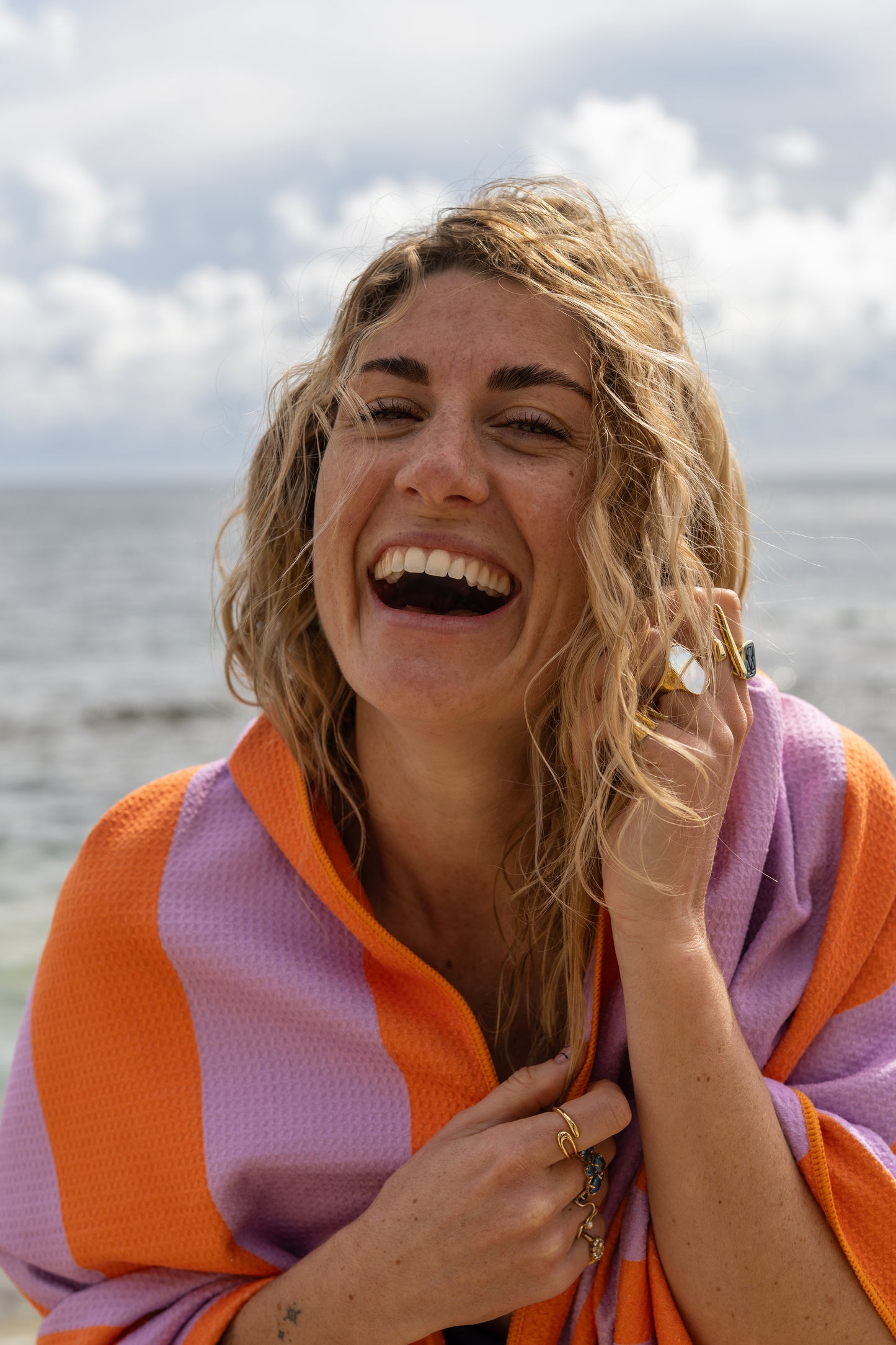 Close-up van een lachende vrouw gewikkeld in het lila-oranje strandlaken van Piece of Sun. Het lichte microvezel met wafelstructuur voelt zacht aan en is gemaakt van 85% gerecycled materiaal.