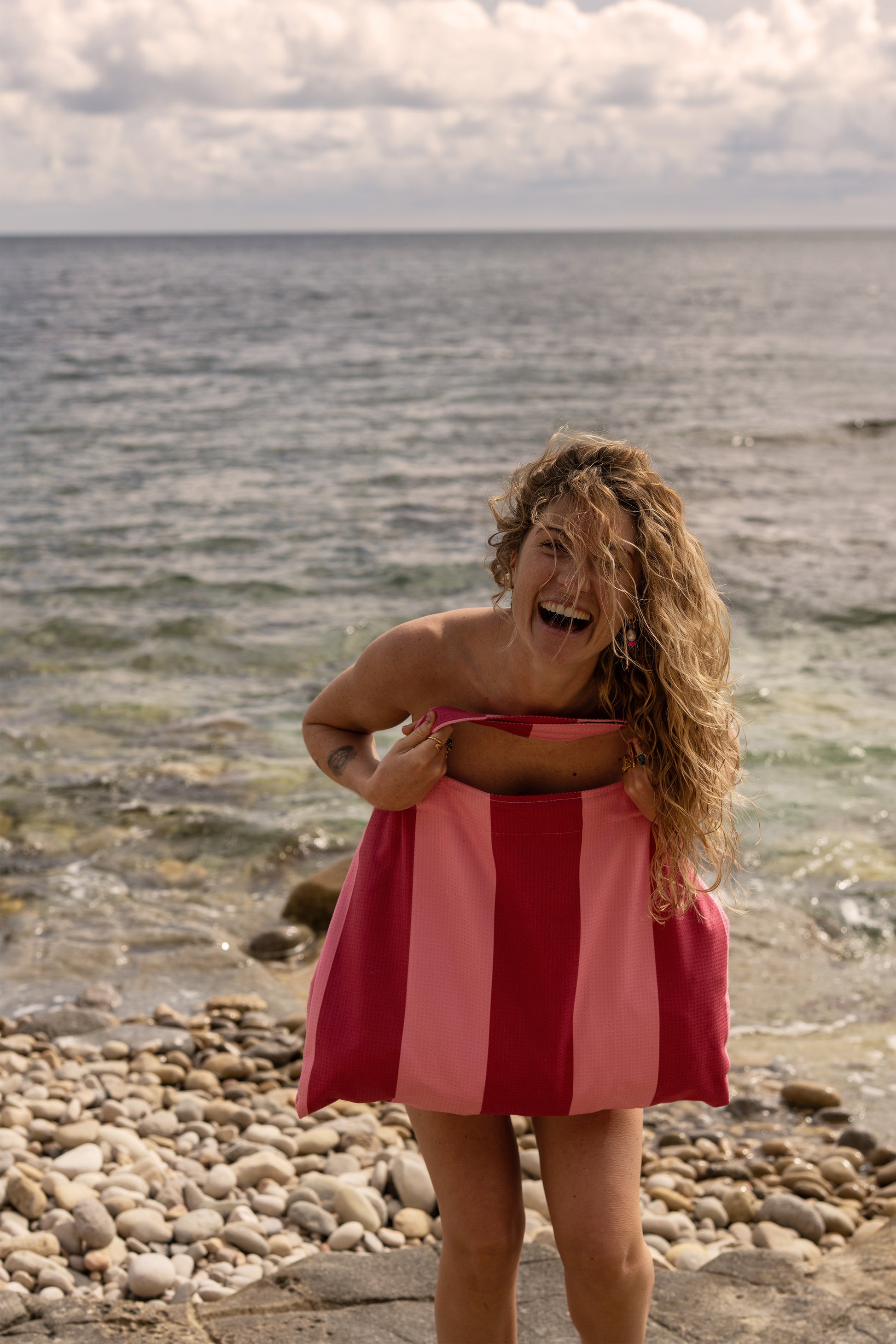 Lachende vrouw bij zee met een gestreepte roze strandtas van Piece of Sun, gemaakt van gerecyclede microvezel – lichtgewicht, compact en ideaal voor stranddagen.