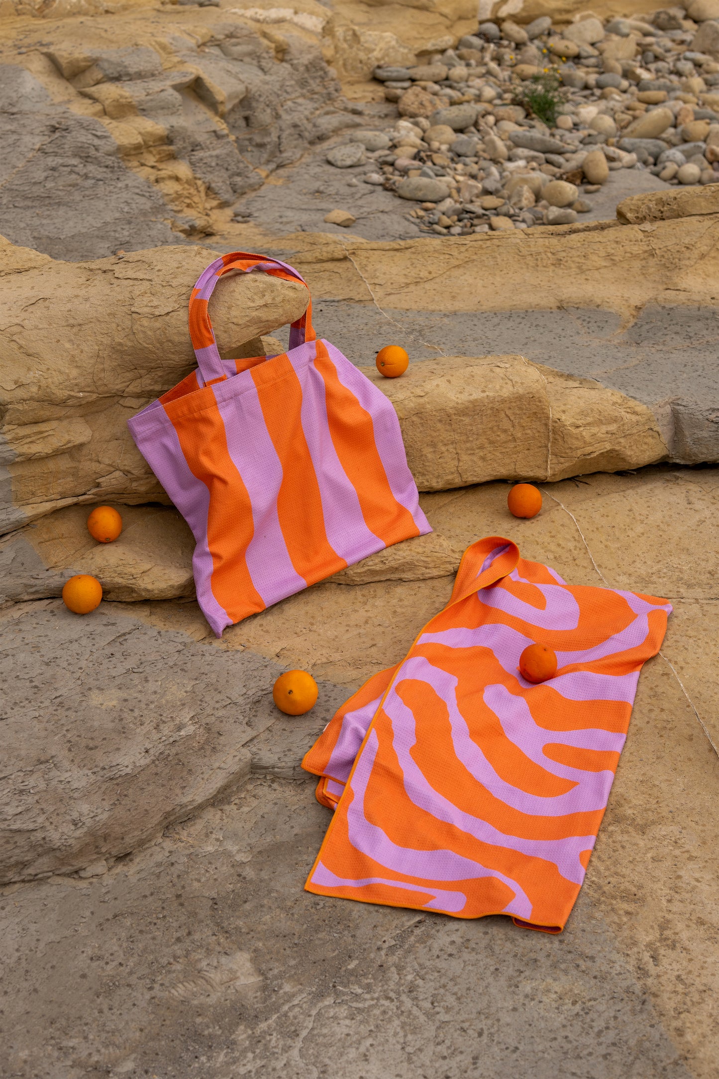 Lila met oranje strandtas en bijpassende zonnehanddoek van Piece of Sun liggen op een zonnige rots tussen sinaasappels. De handdoek is gemaakt van microvezel met wafelstructuur, sneldrogend en voor 85% gerecycled. Perfect voor een stijlvolle dag aan zee of het zwembad.