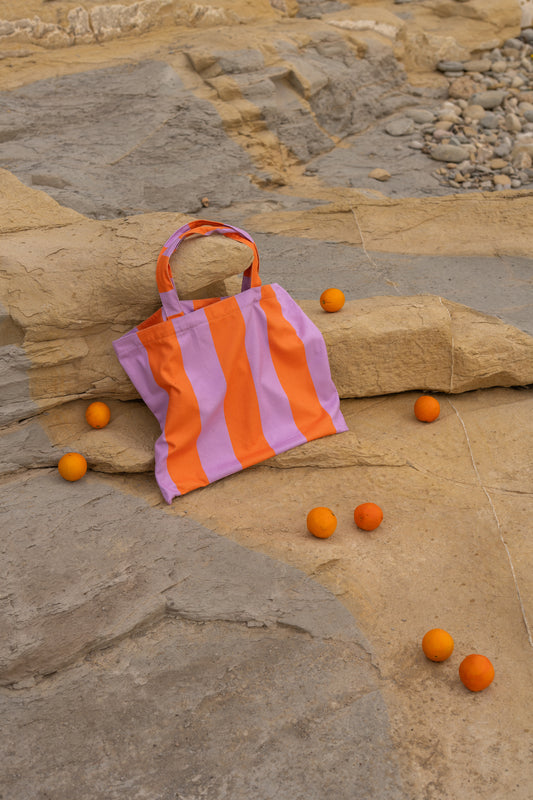 Lila-oranje strandtas van Piece of Sun ligt op een zonnige rots onder verspreide sinaasappels. De tas is gemaakt van microvezel met wafelstructuur, lichtgewicht en ideaal voor stranddagen. Compact opvouwbaar en deels vervaardigd uit gerecycled materiaal.
