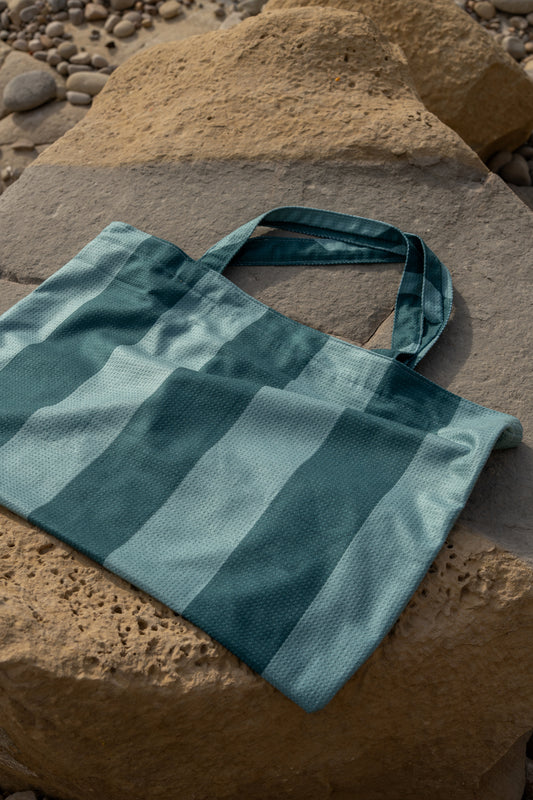 Turquoise met petrol gestreepte strandtas van Piece of Sun, gefotografeerd op een zonnige rots onder natuurlijke lichtval. De tas is gemaakt van duurzame microvezel en past perfect bij de bijbehorende zonnehanddoeken. Ideaal voor een zomerse dag aan het strand.